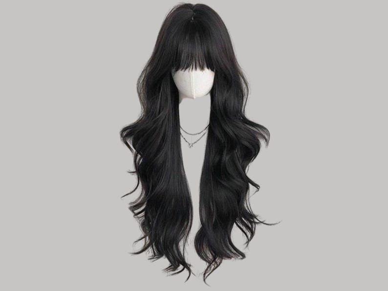 May include: A long, wavy black wig with a blunt fringe. The wig is styled on a mannequin head, and a delicate silver necklace is visible. The hair cascades down in loose waves, creating a natural and voluminous look.