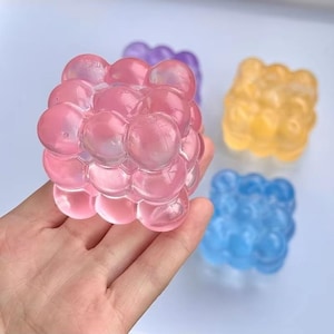 Puede incluir: Una mano sosteniendo un cubo rosa en forma de burbujas. Otros cubos en morado, amarillo y azul son visibles en el fondo. Los cubos están hechos de un material translúcido, lo que les da un aspecto brillante. Los cubos tienen un diseño único y lúdico.
