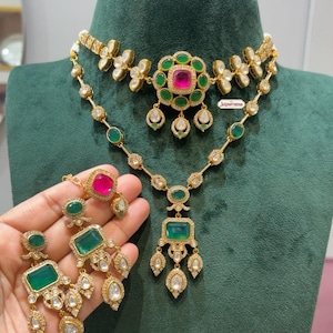 Puede incluir: Un conjunto de collar y pendientes en tono dorado con piedras verde esmeralda y rosa fucsia. El collar presenta un motivo floral central con una piedra rosa, mientras que los pendientes tienen piedras verdes rectangulares y detalles colgantes.