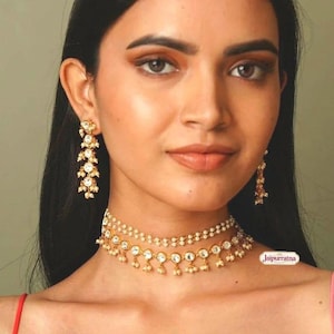 May include: A gold-toned choker necklace and matching drop earrings set featuring white stones and small dangling beads. The necklace consists of two layered chains with circular stone settings and hanging accents. The text Jaipurratna is visible.