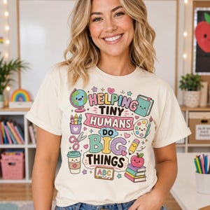 May include: Cream-coloured t-shirt featuring the phrase "Helping Tiny Humans Do Big Things" in a vibrant, playful design. The graphic includes a globe, pencils, scissors, a coffee cup, and an apple on books. The shirt is worn by a person in a classroom.