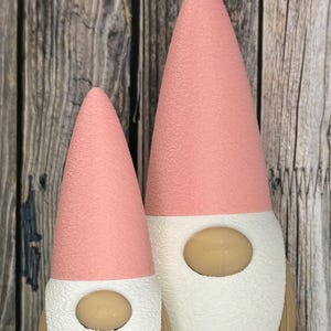 Könnte beinhalten: Zwei dekorative Zwerge mit rosa konischen Hüten, weißen Bärten und beigen Nasen. Die Zwerge haben einen strukturierten, gerippten Sockel und stehen vor einem Hintergrund aus Holzplanken. Ein Zwerg ist kleiner als der andere.