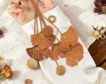 May include: A key ring with a brown leather strap and decorative leaf-shaped charms. The charms are in shades of orange and brown, with a textured appearance. The key ring is held against a white shirt sleeve.
