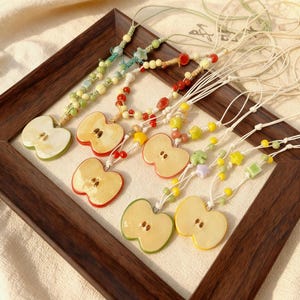 May include: A collection of handmade necklaces featuring ceramic apple slice pendants. Each pendant is a different color, including green, red, and yellow, and is strung on a cord with colorful beads. The necklaces are displayed on a wooden frame.