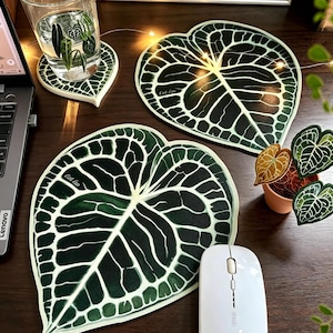 Puede incluir: Un juego de posavasos con forma de hoja y un alfombrilla de ratón a juego. Los posavasos y la alfombrilla presentan un diseño de hoja verde oscuro con vetas blancas. Un vaso de agua con un diseño de hoja está sobre uno de los posavasos. También hay un ratón de ordenador blanco.