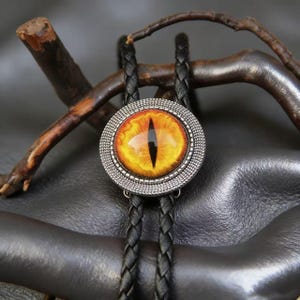 Puede incluir: Una corbata bolo con un colgante redondo que representa un ojo amarillo y naranja detallado. El ojo está engastado en un marco plateado texturizado. La corbata tiene un cordón de cuero trenzado negro.