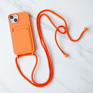 May include: Bright orange phone case with a cardholder and matching lanyard. The case has a smooth finish and is designed to hold a phone. The lanyard is a thick, woven cord with adjustable toggles.