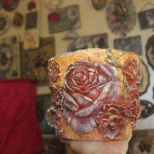 May include: A gold and red decorative planter with a raised design of roses. The planter is made of a textured material and has a rustic look.