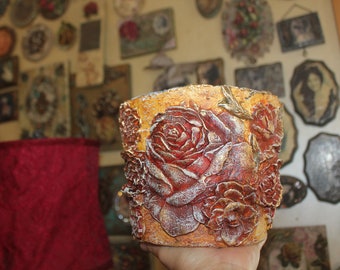 May include: A gold and red decorative planter with a raised design of roses. The planter is made of a textured material and has a rustic look.