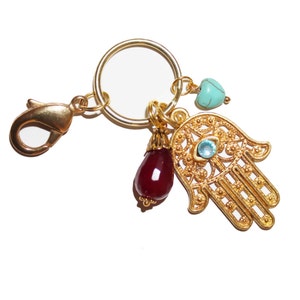 Puede incluir: Llavero dorado con un dije de mano de hamsa, una cuenta roja en forma de lágrima y un dije de corazón turquesa. El dije de mano de hamsa tiene un detalle de ojo malvado.