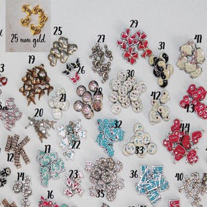 May include: A collection of small metal charms and buttons arranged in numbered groups, featuring various shapes including hearts, bows, flowers, and symbols in colors like red, blue, pink, gold, and silver, with some items containing rhinestones.