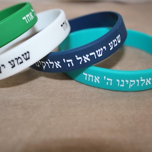 May include: A collection of colorful silicone wristbands. The bands are in shades of green, white, teal, and navy blue. Each band has white Hebrew text. The bands are stacked on top of each other.