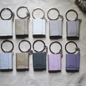 May include: Twelve small leather keychains with silver key rings and chains. The keychains are in various colours, including white, grey, pink, purple, and brown.