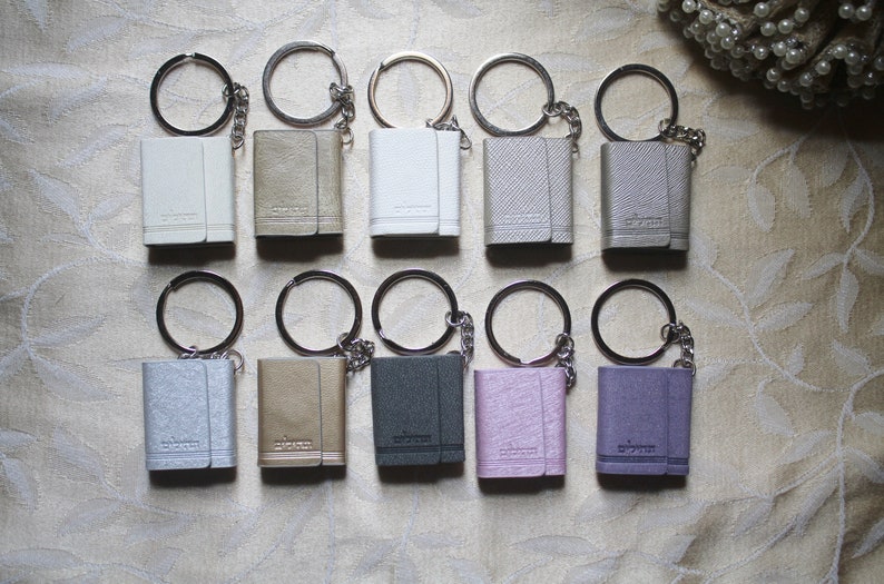May include: Twelve small leather keychains with silver key rings and chains. The keychains are in various colours, including white, grey, pink, purple, and brown.