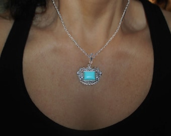 Puede incluir: Un collar de cadena de plata con un colgante de piedra turquesa. El colgante está en un marco decorativo y adornado.