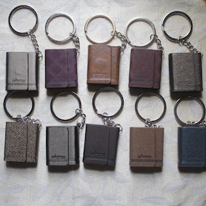 May include: Ten leather keychains with silver key rings. Each keychain is a small, rectangular book-shaped leather case with a silver metal clasp. The keychains are arranged in two rows of five.