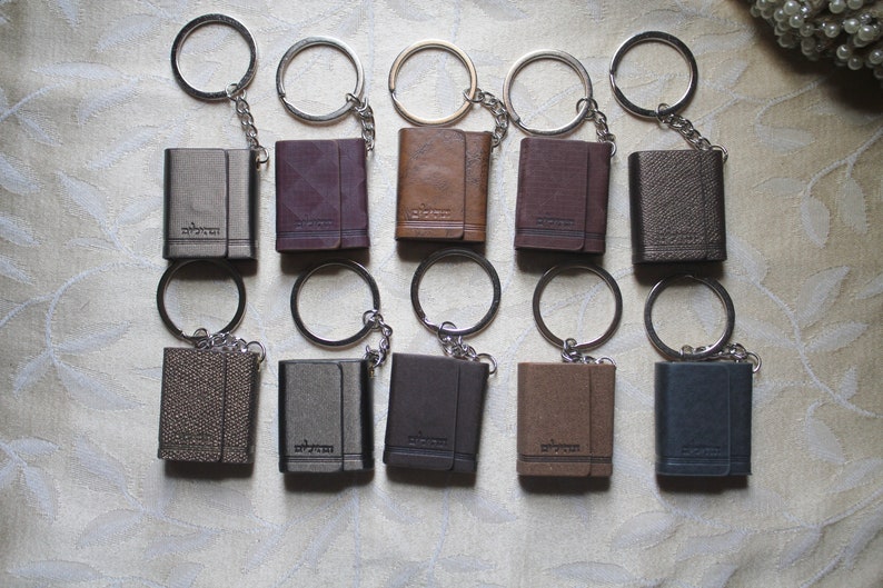 May include: Ten leather keychains with silver key rings. Each keychain is a small, rectangular book-shaped leather case with a silver metal clasp. The keychains are arranged in two rows of five.