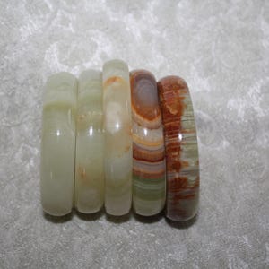 May include: A set of five smooth, polished stone rings arranged in a row. The rings feature varying natural patterns and colors, ranging from pale translucent green and cream to banded shades of orange, brown, and deep green with visible mineral swirls.
