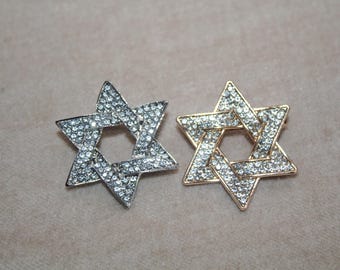May include: Two Star of David brooches. One is silver-toned, the other gold-toned. Both are encrusted with small, clear crystals. The brooches are set against a neutral background.