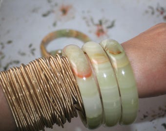 May include: A collection of jewelry featuring a large stack of thin, metallic gold-toned wire bangles worn alongside three smooth, polished, pale green stone bangle bracelets with subtle orange and white banding patterns.