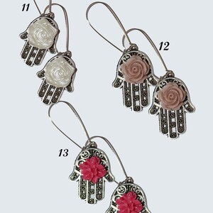 以下が含まれることがあります： 異なる色の花のアクセントが付いた、銀色のハmsaハンドイヤリングの3ペア。最初のペアは白い花、2番目のペアはピンクの花、3番目のペアは赤い花です。