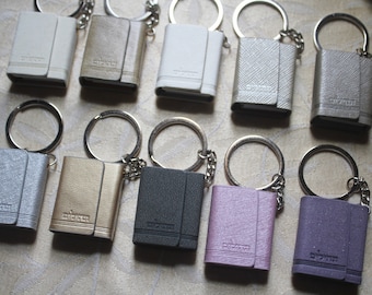 Könnte beinhalten: Zehn kleine, ledergebundene Schlüsselanhänger mit silbernen Schlüsselringen. Die Schlüsselanhänger sind in verschiedenen Farben, darunter Silber, Gold, Schwarz, Weiß, Rosa und Lila. Jeder Schlüsselanhänger hat das Wort "Sticker" auf der Vorderseite geprägt.
