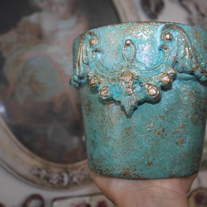 May include: A turquoise-colored, textured ceramic pot with ornate gold-colored embellishments. The pot has a distressed finish and a decorative design around the rim. A unique piece for home decor.