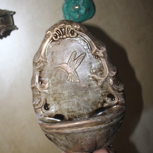 Puede incluir: Una pieza de decoración de pared de cerámica con un acabado dorado y marrón. La decoración presenta un diseño en relieve de un colibrí en vuelo. La pieza tiene un marco decorativo y ornamentado.