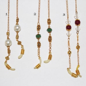 May include: Three gold eyeglass chains with different colored beads. The first chain has white pearls, the second has green beads, and the third has red and clear beads.