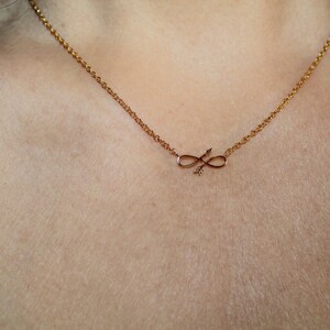 May include: A gold chain necklace with a delicate infinity symbol pendant featuring an arrow design.