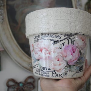 May include: A decorative ceramic pot with a textured, off-white rim and a floral design. The pot features pink and white peonies and text that reads "Paris". The pot is held in a hand.