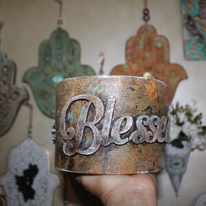 May include: A rustic, copper-colored metal container with the word "Blessed" embossed in silver lettering. The container has a distressed, aged look with a turquoise and gold paint finish.