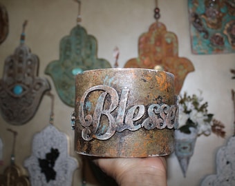 May include: A rustic, copper-coloured metal container with the word "Blessed" embossed in silver lettering. The container has a distressed, aged look with a turquoise and gold paint finish.