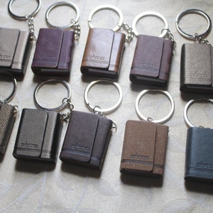 May include: Ten small leather-bound notebooks with keychains. The notebooks are in various colours, including brown, black, grey, and gold. Each notebook has a silver keychain attached to it.