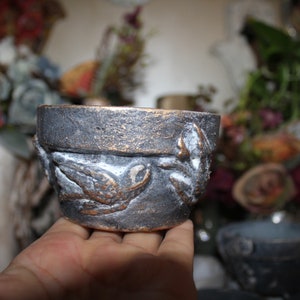 Puede incluir: Un pequeño macetero de cerámica gris oscuro con un acabado metálico dorado. El macetero tiene un diseño en relieve de pájaros y hojas.