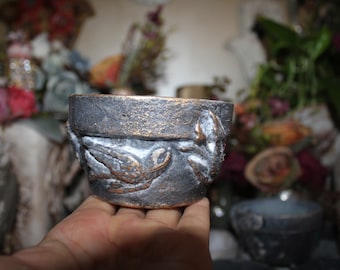 Puede incluir: Un pequeño macetero de cerámica gris oscuro con un acabado metálico dorado. El macetero tiene un diseño en relieve de pájaros y hojas.