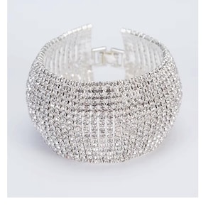 May include: A silver bangle bracelet with a wide, rounded design. The bracelet is completely covered in small, clear rhinestones.