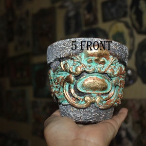 Puede incluir: Un macetero de cerámica gris con un diseño en color turquesa y dorado. El macetero tiene un diseño decorativo y ornamentado con una cara de calavera en el centro. El texto "5 FRONT" es visible en el macetero.