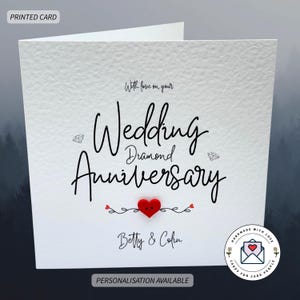 May include: A white card for a wedding anniversary, featuring the words "Wedding Diamond Anniversary" in black script. A red heart button is centered below the text, with the names "Betty & Colin" also included.
