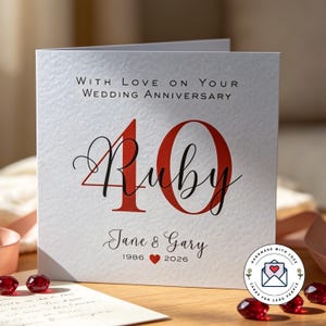 May include: A square white greeting card with the text "WITH LOVE ON YOUR WEDDING ANNIVERSARY" at the top. The number "40" is printed in large red font with the word "Ruby" in black script overlaying it. Below, "Jane & Gary" appears with "1986", a red heart, and "2026".