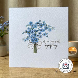 May include: A square white card featuring a watercolour bouquet of blue forget-me-nots, tied with a brown ribbon. The card has the text "With Love and Sympathy" in black script. A small sticker with a heart is in the bottom right corner.