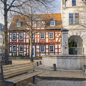 May include: A view of a European town square with a wooden bench, cobblestone paving, and historic buildings. The buildings have red tile roofs and white and red timber-framed walls. A stone fountain is also visible.