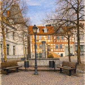 May include: A pencil drawing of a European town square. The scene features a cobbled plaza with benches, a fountain, and a vintage street lamp. Buildings with red tile roofs and timber-framed walls line the square under a blue sky.