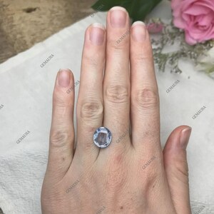May include: A light blue, oval-cut gemstone. The gemstone is set on a hand, showcasing its color and clarity. The background includes a white cloth and a pink rose, providing a soft contrast to the gem's cool tones. The gemstone appears to be approximately 1 inch in length.