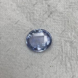 May include: A faceted, oval-shaped, light blue gemstone. The gem has a polished surface with multiple facets that reflect light. The stone is set against a neutral, textured background, highlighting its color and clarity. This gemstone is likely for jewelry.