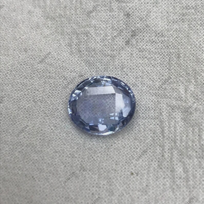 May include: A faceted, oval-shaped, light blue gemstone. The gem has a polished surface with multiple facets that reflect light. The stone is set against a neutral, textured background, highlighting its color and clarity. This gemstone is likely for jewelry.