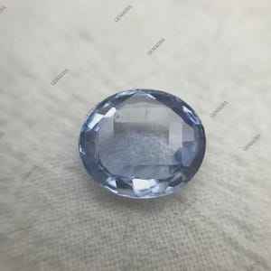 May include: A faceted, oval-shaped, light blue gemstone. The gem has a polished surface with multiple facets that reflect light. The gemstone is set against a white background. The image has the watermark "GEMAURA" repeated around the gem.