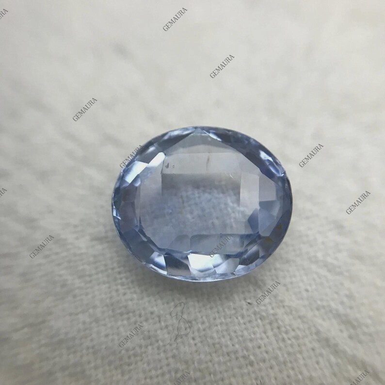 May include: A faceted, oval-shaped, light blue gemstone. The gem has a polished surface with multiple facets that reflect light. The gemstone is set against a white background. The image has the watermark "GEMAURA" repeated around the gem.
