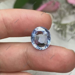May include: An oval-cut, light blue gemstone with multiple facets. The gem is held between fingers, showcasing its clarity and color. The stone reflects light, creating a sparkling effect. The background is blurred, focusing attention on the gemstone.