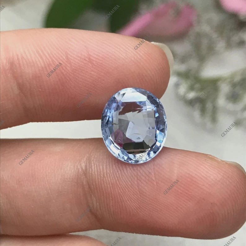 May include: An oval-cut, light blue gemstone with multiple facets. The gem is held between fingers, showcasing its clarity and color. The stone reflects light, creating a sparkling effect. The background is blurred, focusing attention on the gemstone.